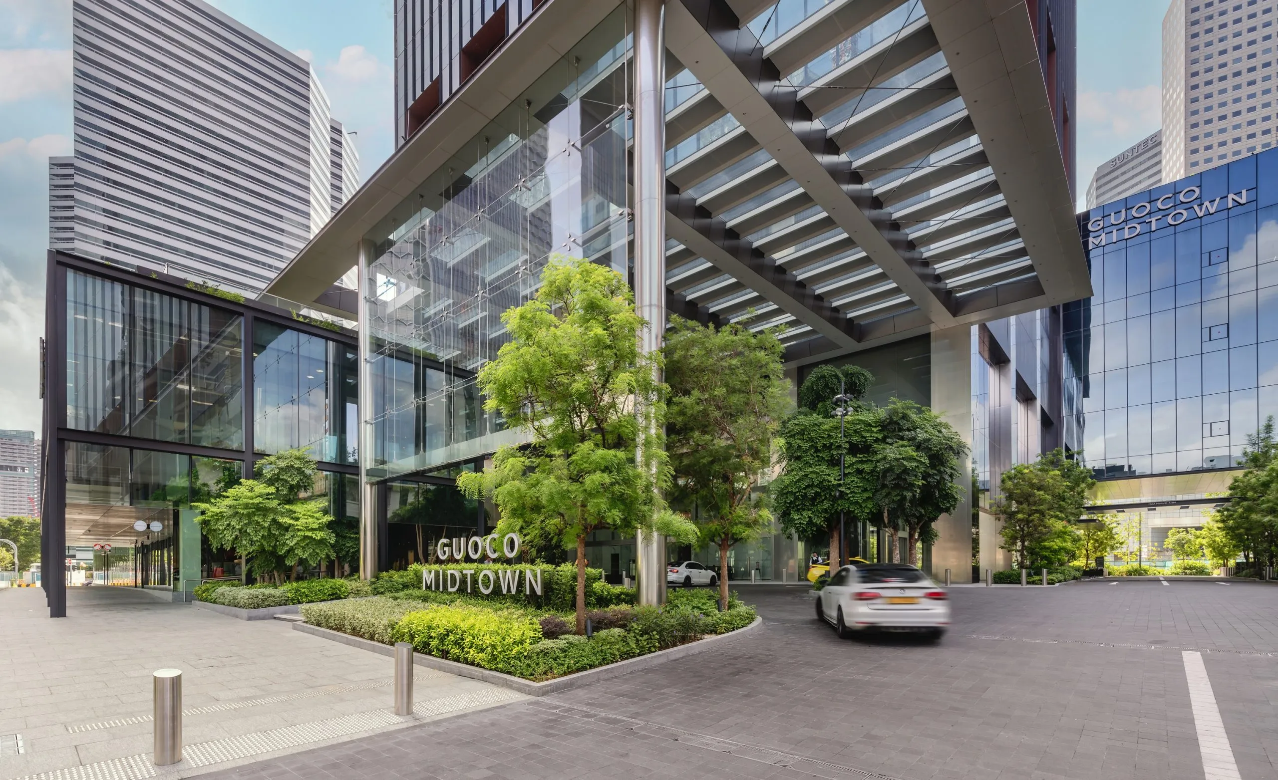 Modern glass and steel entrance of Guoco Midtown building with greenery and cars driving in front.