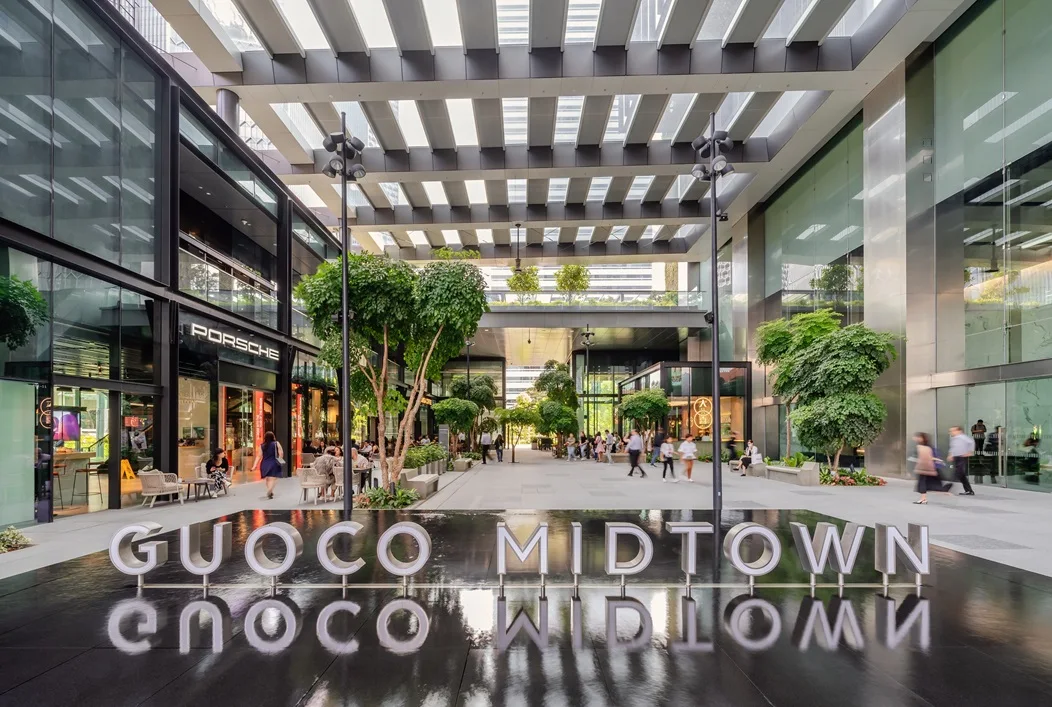 Modern open-air Guoco Midtown shopping area with glass buildings, green trees, and a reflective sign spelling Guoco Midtown.
