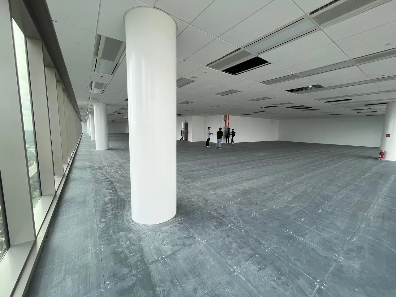 Spacious empty office space with large white columns, floor-to-ceiling windows on the left, and a group of people standing near the back wall.