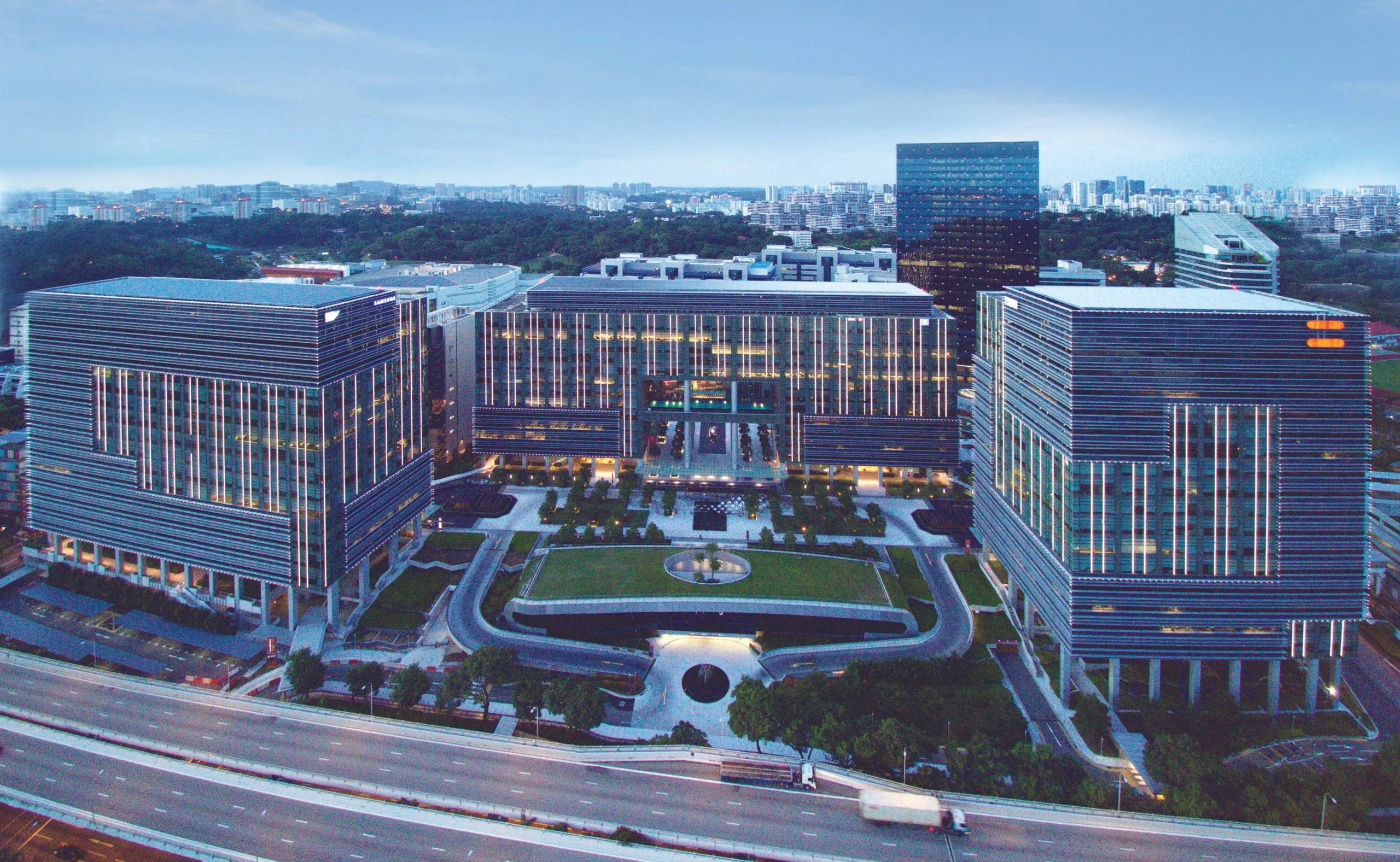 Aerial view of a Mapletree Business City with three large office buildings and landscaped grounds, set beside a multi-lane highway.